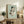 May include: Framed reproduction of early 20th century figurative artwork showing a crouching nude figure rendered in muted earth tones against pale green background, displayed on rustic wooden sideboard alongside fern plant, ceramic vase, and stacked books in warmly lit interior setting with cream upholstered furniture.