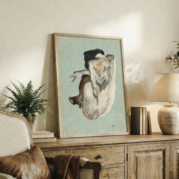 May include: Framed reproduction of early 20th century figurative artwork showing a crouching nude figure rendered in muted earth tones against pale green background, displayed on rustic wooden sideboard alongside fern plant, ceramic vase, and stacked books in warmly lit interior setting with cream upholstered furniture.