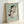 May include: Framed reproduction of an early 20th century expressionist drawing showing a nude female figure in crouching pose with dark hair, rendered in muted watercolor tones of brown, blue and flesh against pale green background. Natural wood frame displayed leaning against white wall in soft natural lighting.