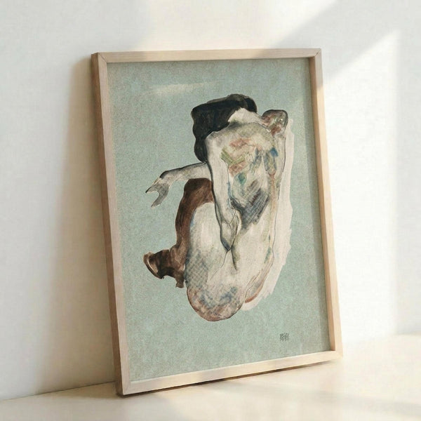 May include: Framed reproduction of an early 20th century expressionist drawing showing a nude female figure in crouching pose with dark hair, rendered in muted watercolor tones of brown, blue and flesh against pale green background. Natural wood frame displayed leaning against white wall in soft natural lighting.