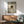 May include: Framed poster print showing a black cat with bright yellow eyes reading a small book while tucked under green and white striped bedding. The artwork is displayed above a wooden sideboard with a white ceramic table lamp in a modern living room with sage green upholstered chair and round coffee table. Warm sunlight streams across white walls creating geometric shadows.