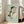 May include: Framed reproduction of expressionist figure drawing showing crouching nude woman rendered in muted earth tones and pale greens on sage background, displayed in light wood frame leaning against white wall beside beige upholstered chair in bright modern living room with large windows and hardwood floors.