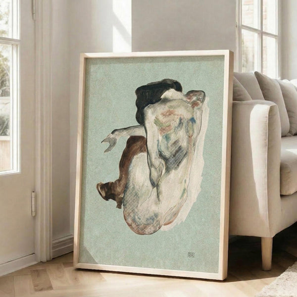 May include: Framed reproduction of expressionist figure drawing showing crouching nude woman rendered in muted earth tones and pale greens on sage background, displayed in light wood frame leaning against white wall beside beige upholstered chair in bright modern living room with large windows and hardwood floors.