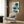 May include: Framed botanical artwork featuring blue poppies with Oriental calligraphy displayed on wood-paneled wall above beige upholstered armchair with geometric patterned pillow. Modern brass floor lamp and dark round side table with open book complete the neutral-toned interior setting with natural lighting from window.