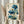May include: hands holding a framed botanical print featuring vibrant blue poppies with stems, leaves, and seed pods against aged beige paper. Asian calligraphy text appears on the left side with red seal stamps. Person wears cream-colored ruffled sleeves while displaying the artwork near dried pampas grass and white curtains in soft natural lighting.