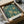 May include: Framed William Morris floral pattern print in teal and gold, featuring ornate chrysanthemums and decorative foliage, displayed in black frame inside open cardboard shipping box with tissue paper wrapping, surrounded by natural lighting on neutral surface.