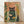 May include: Framed poster print leaning against white wall on light wood floor with sunlight shadows. Shows whimsical illustration of black cat wearing pink glasses perched on rim of large glass jar containing three orange goldfish swimming among green aquatic plants. Background features coral-pink tones with botanical foliage in greens and yellows. Natural wood frame, approximately 16x20 inch scale.