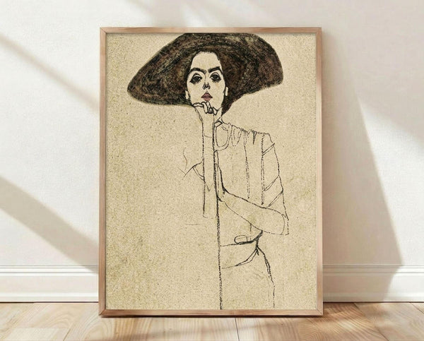 May include: Framed art print leaning against white wall on light wooden floor. The poster features a line drawing of a woman with voluminous dark hair wearing a large hat, her hand positioned near her face. The artwork is rendered in black ink on cream-colored textured paper, displaying an early 20th century expressionist style. Natural light casts soft shadows across the scene, and the simple wooden frame complements the vintage aesthetic of the illustration.