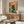May include: Framed artwork showing a black cat wearing pink glasses peering over a fishbowl containing orange goldfish, surrounded by green foliage on a coral background. Displayed above a wooden credenza with sliding doors in a modern living room with white curved chair, glass coffee table, and warm natural lighting.