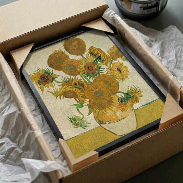 May include: Framed reproduction of Van Gogh's sunflowers painting showing vibrant yellow blooms in a vase, displayed in a black frame being unpacked from a cardboard shipping box with protective wrapping paper and wooden corner guards on a white surface
