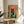 May include: Framed art print displayed on round wooden side table next to beige upholstered chair. The colorful illustration shows a black cat wearing pink glasses perched behind a fishbowl containing three orange goldfish and aquatic plants, surrounded by green foliage on a coral pink background. Natural sunlight streams across the neutral-toned room setting with houseplant visible in corner.