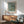 May include: Framed Van Gogh almond blossom poster with blue background and white flowering branches displayed above wood sideboard. Mid-century modern room setting with mint green upholstered armchair, white ceramic table lamp, and natural sunlight casting shadows on white walls.