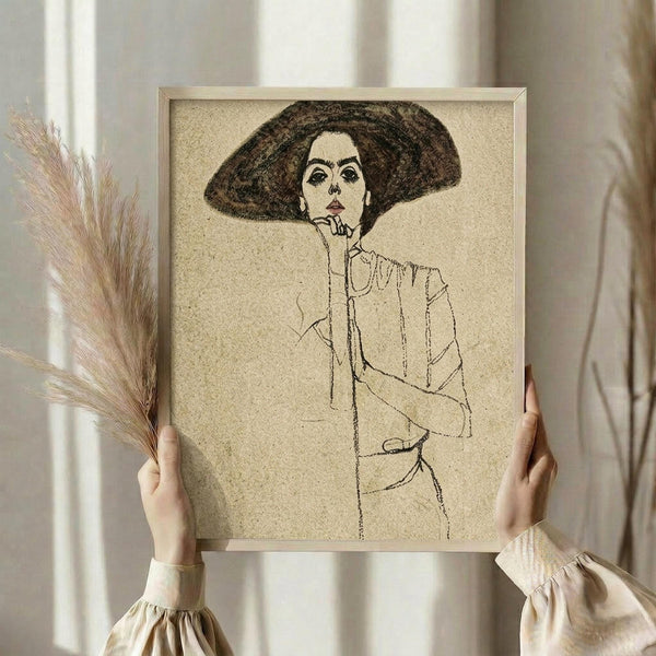 May include: Person in cream ruffled blouse holding framed art print showing minimalist line drawing of woman in wide-brimmed hat, rendered in brown tones on textured beige background. Light wooden frame, soft natural lighting, dried wheat stems visible, neutral interior setting.
