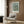 May include: Van Gogh's Almond Blossom artwork poster with blue background and white flowering branches, displayed in light wooden frame on wood-paneled wall. Modern beige armchair with geometric patterned pillow sits below, alongside brass floor lamp and dark side table with open book. Neutral tones throughout room setting with natural lighting from window.