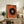 May include: Abstract geometric poster print in warm wood frame, featuring large black circle with colorful center rings on textured orange background with radiating lines. Displayed on rustic wooden sideboard alongside potted fern, ceramic vases, and books in cozy living room setting with neutral walls and brown leather furniture.