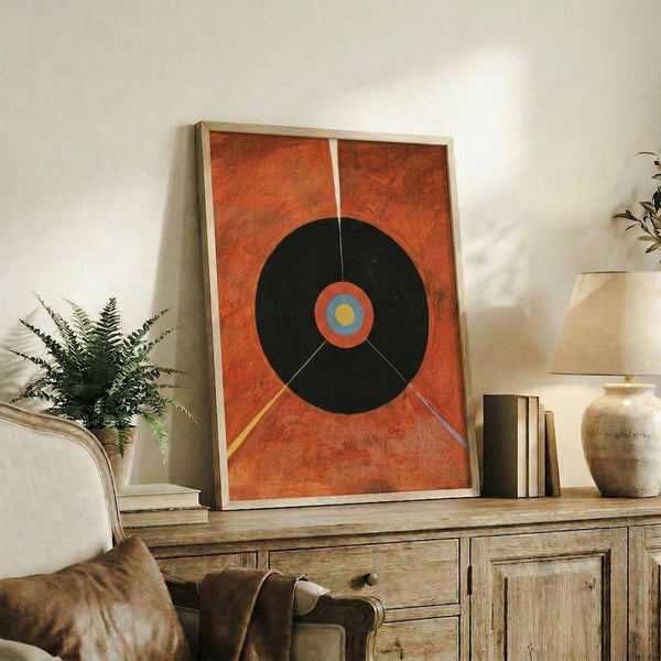 May include: Abstract geometric poster print in warm wood frame, featuring large black circle with colorful center rings on textured orange background with radiating lines. Displayed on rustic wooden sideboard alongside potted fern, ceramic vases, and books in cozy living room setting with neutral walls and brown leather furniture.
