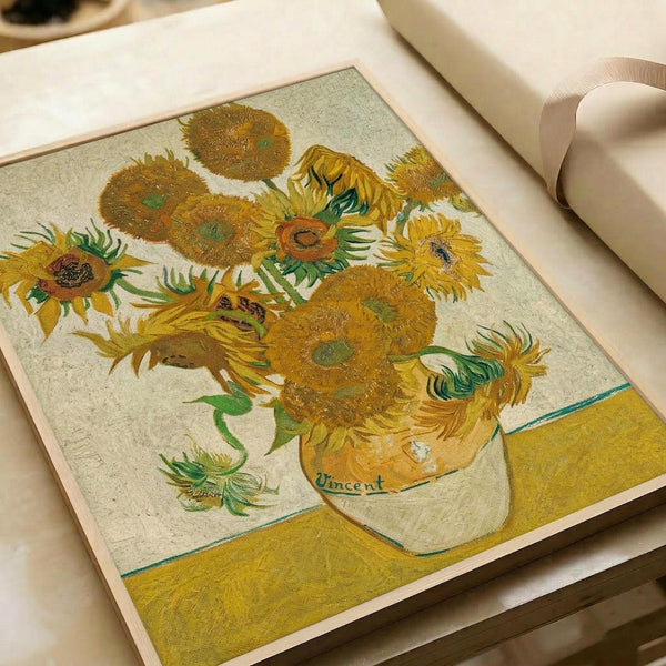 May include: Van Gogh's iconic sunflowers painting print displayed on a cream-colored upholstered chair or sofa. The artwork features vibrant yellow sunflowers in various stages of bloom arranged in a ceramic vase against a pale background with yellow table surface. Vincent's signature is visible on the vase. The print has a light wooden or natural frame and shows the characteristic thick brushstrokes and warm golden tones of the original oil painting.