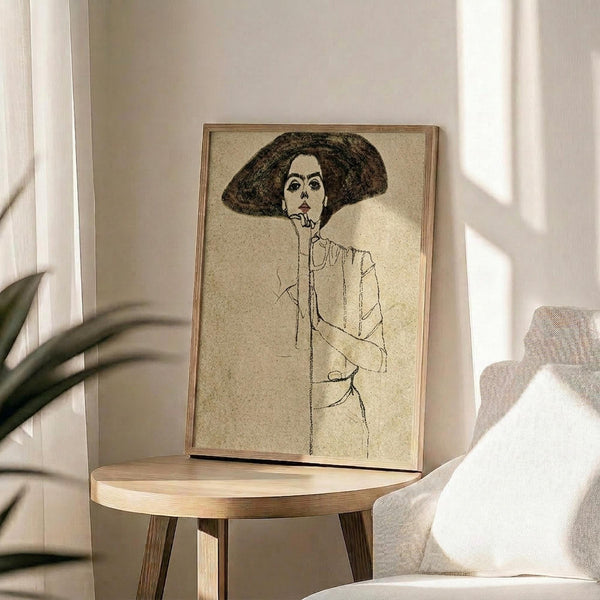 May include: Framed sketch print of a woman wearing a large dark hat and white blouse, displayed on a round wooden side table next to a beige upholstered chair. Natural sunlight streams across the neutral-toned room setting with plant leaves visible in the corner. The artwork features minimal line drawing style with earth tones against cream background in a thin wooden frame.