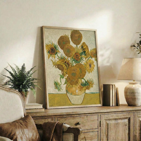 May include: Van Gogh's Sunflowers painting reproduction in light wood frame, displayed on rustic wooden sideboard alongside fern plant, books, and ceramic vase. Warm living room setting with beige chair and neutral wall backdrop. The artwork features golden yellow sunflowers in white vase against yellow surface with textured brushstroke style characteristic of post-impressionist art.