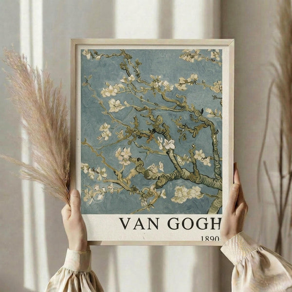 May include: Person in beige clothing with ruffled sleeves holding framed Van Gogh almond blossom print showing delicate white flowers on twisting branches against soft blue background. Light wood frame with white matting displays "VAN GOGH 1890" text below artwork. Dried pampas grass stems visible on left side against neutral curtained window background.