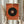May include: Person holding framed abstract art print featuring large black circle on textured orange background with concentric circles in center (orange, blue, yellow) and geometric lines extending outward. Modern minimalist artwork in light wooden frame, displayed in neutral interior setting with dried wheat stems and white curtains.