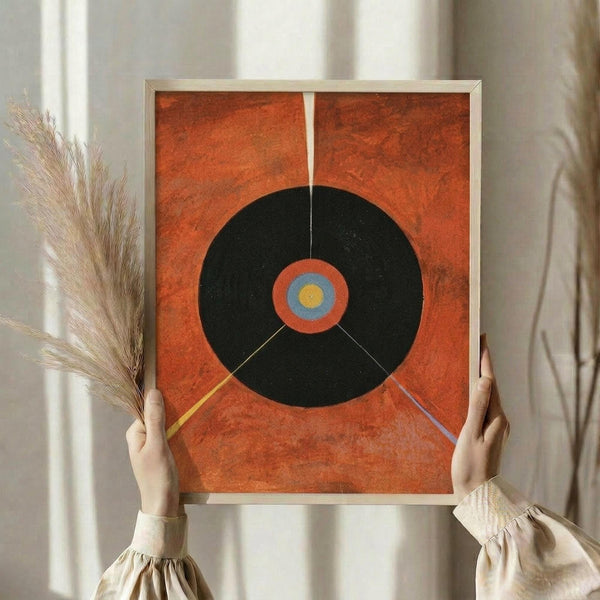 May include: Person holding framed abstract art print featuring large black circle on textured orange background with concentric circles in center (orange, blue, yellow) and geometric lines extending outward. Modern minimalist artwork in light wooden frame, displayed in neutral interior setting with dried wheat stems and white curtains.
