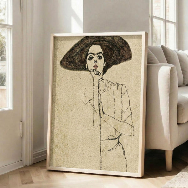 May include: Framed art print leaning against white wall next to beige upholstered chair in bright living room with hardwood floors and windows. The artwork shows a sketched portrait of a woman wearing a large dark hat, drawn in black lines on cream textured background with minimalist style. Light wood frame surrounds the vintage-inspired illustration.