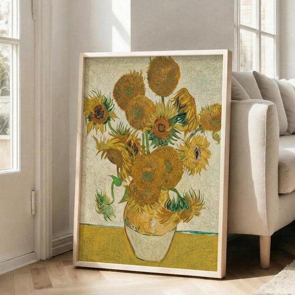 May include: Framed reproduction of Van Gogh's famous sunflowers painting leaning against white wall in bright living room. Yellow and orange sunflowers in ceramic vase against pale yellow background, displayed in light wood frame. Gray upholstered sofa and large windows with natural light visible in modern interior setting.