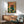 May include: Framed poster print hanging on white wall above wooden sideboard, showing black cat wearing pink glasses peering into fishbowl with orange goldfish and green aquatic plants, surrounded by botanical foliage on coral background. Mid-century modern living room with sage green upholstered chair, small white table lamp, and natural sunlight creating shadows across the scene.