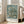 May include: Framed reproduction of Van Gogh's almond blossom painting featuring white flowers on twisted brown branches against a turquoise blue background, displayed on light wooden floors next to a beige upholstered sofa in a bright room with white walls and large windows. The poster has "VAN GOGH 1890" text at the bottom and appears medium-sized in a natural wood frame.