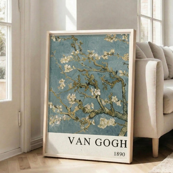 May include: Framed reproduction of Van Gogh's almond blossom painting featuring white flowers on twisted brown branches against a turquoise blue background, displayed on light wooden floors next to a beige upholstered sofa in a bright room with white walls and large windows. The poster has "VAN GOGH 1890" text at the bottom and appears medium-sized in a natural wood frame.