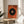 May include: Abstract geometric poster in thin wood frame leaning against white wall on round wooden side table. Orange textured background with large black circle containing concentric rings in orange, blue and yellow center. Colorful radiating lines extend from circle. Modern living room setting with beige upholstered chair and plant shadow visible.