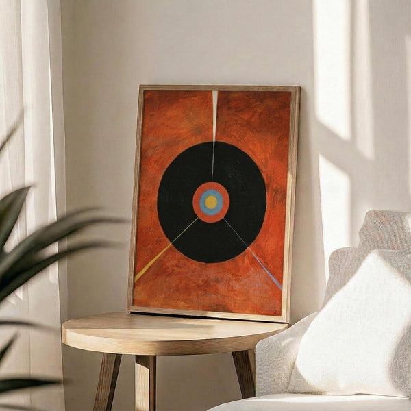 May include: Abstract geometric poster in thin wood frame leaning against white wall on round wooden side table. Orange textured background with large black circle containing concentric rings in orange, blue and yellow center. Colorful radiating lines extend from circle. Modern living room setting with beige upholstered chair and plant shadow visible.