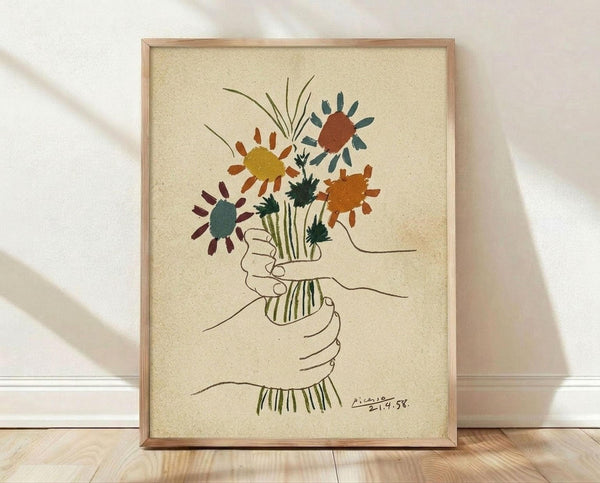 May include: Framed art print featuring a line drawing of hands holding a bouquet of stylized flowers in earth tones - yellow, orange, rust, and teal blooms with green stems. The minimalist artwork has a cream textured background, thin wooden frame, and signature marking. Displayed against white fabric backdrop on light wooden floor with natural shadows.