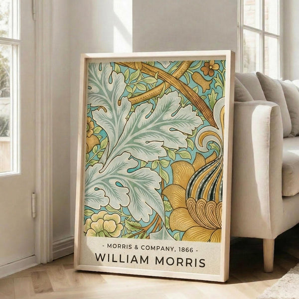 May include: Large framed botanical poster featuring William Morris's ornate floral pattern in sage green, golden yellow, and turquoise. The design shows intricate acanthus leaves, flowing vines, and decorative flowers in the Arts and Crafts style. The print is displayed in a light wooden frame leaning against white walls in a bright living room with beige armchair and large windows.