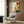 May include: Colorful botanical poster with vibrant sunburst flowers in red, yellow, orange and pink tones displayed above a beige upholstered armchair with geometric pillow. Modern floor lamp with brass finish stands nearby. Wood-paneled wall background with natural lighting from window curtains creates warm, contemporary living room setting.