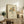 May include: Framed artwork showing colorful abstract flowers in a hand-drawn bouquet with line art hands holding stems, displayed on a rustic wooden credenza alongside a fern plant, books, and ceramic vase in a warm, neutral-toned living room setting with beige furniture.