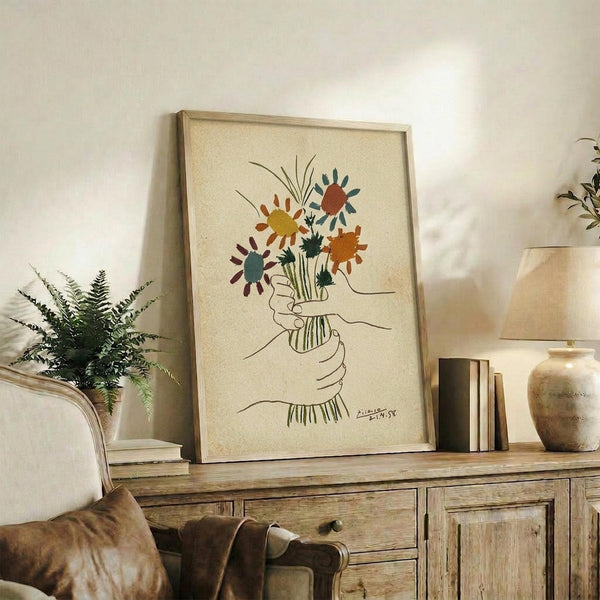 May include: Framed artwork showing colorful abstract flowers in a hand-drawn bouquet with line art hands holding stems, displayed on a rustic wooden credenza alongside a fern plant, books, and ceramic vase in a warm, neutral-toned living room setting with beige furniture.