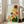 May include: Large framed botanical artwork leaning against white wall next to beige sofa. The vertical print features colorful stylized flowers in yellow, orange, red, and pink with radiating petal patterns resembling fireworks or dandelions. Green stems and leaves fill the bottom portion against a cream background. Natural light from nearby windows illuminates the bright, cheerful floral design in this modern living room setting.