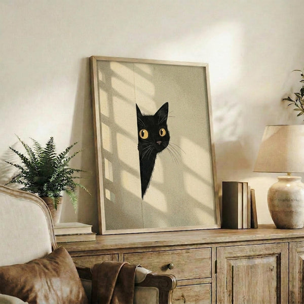 May include: Framed artwork featuring a black cat with bright golden eyes peeking out, displayed on a rustic wooden sideboard alongside books, a ceramic vase, and a fern plant. Natural sunlight creates diagonal shadows across the beige textured background of the minimalist artwork. The wooden frame complements the warm, earthy home decor setting.