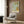 May include: Framed botanical art print with ornate green and gold foliage design hanging on wood-paneled wall above beige upholstered armchair with geometric throw pillow. Modern brass floor lamp and round black side table with open book complete the elegant reading nook scene with neutral curtains and warm lighting.