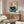 May include: Framed vintage-style poster featuring a turquoise record player with vinyl record and Japanese characters on cream background, displayed above mid-century wooden credenza with sliding doors. White curved chair and glass coffee table with colorful abstract design visible in modern living room setting with white walls and natural lighting.