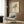 May include: A framed art print featuring colorful abstract flowers in a minimalist line drawing style, displayed on a wood-paneled wall above a beige upholstered armchair with geometric patterned pillow. The artwork shows vibrant orange, yellow, and teal blooms with delicate black line work depicting hands holding the bouquet. Modern brass floor lamp and dark round side table with open book complete the warm, contemporary living space setting.