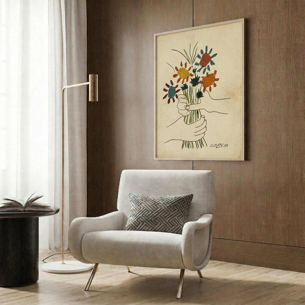 May include: A framed art print featuring colorful abstract flowers in a minimalist line drawing style, displayed on a wood-paneled wall above a beige upholstered armchair with geometric patterned pillow. The artwork shows vibrant orange, yellow, and teal blooms with delicate black line work depicting hands holding the bouquet. Modern brass floor lamp and dark round side table with open book complete the warm, contemporary living space setting.