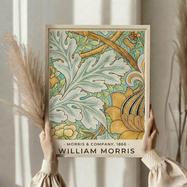 May include: Person in cream ruffled sleeves holding framed botanical art print featuring ornate foliage pattern in sage green, golden yellow, and turquoise blue. Victorian-style decorative design with flowing acanthus leaves and stylized flowers in light wooden frame. Dried pampas grass visible on left side against soft neutral curtained background.