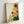 May include: Framed botanical art print leaning against white wall, featuring stylized flowers in warm tones - red, orange, yellow, and pink circular blooms with radiating線patterns resembling dandelions or sunbursts, accompanied by green foliage on cream background, displayed in natural wood frame