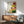 May include: Framed botanical print featuring colorful sunburst flowers in red, yellow, orange, and pink with green foliage, displayed above a mid-century modern wooden credenza with table lamp in a bright living room setting with sage green upholstered chair and round coffee table