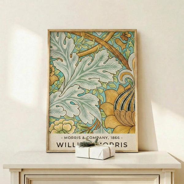 May include: Framed botanical art print featuring ornate acanthus leaves and flowers in sage green, golden yellow, and turquoise colors with Victorian-style decorative flourishes. The print displays "Morris & Company, 1866" and "William Morris" text at bottom. Shown in thin natural wood frame on light wooden shelf against cream wall, with small white wrapped gift box placed in front.
