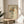 May include: Framed art print displaying a line drawing of hands holding a colorful bouquet with flowers in orange, yellow, teal, and burgundy tones on cream paper. The wooden-framed artwork sits on a round natural wood side table next to a beige upholstered chair with white pillow. Soft natural lighting creates shadows on the neutral wall, with green plant leaves visible in the corner of the modern, minimalist room setting.