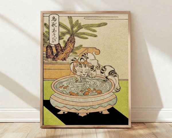 May include: Framed Japanese woodblock print showing a striped tabby cat drinking from an ornate ceramic bowl with floating fish, set on a green tatami mat. Palm trees visible in background with Japanese text in upper left corner. Print displayed in light wood frame against white wall on wooden floor.